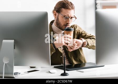Selective focus of 3d artist looking at wristwatch while drinking coffee near digital devices on table Stock Photo