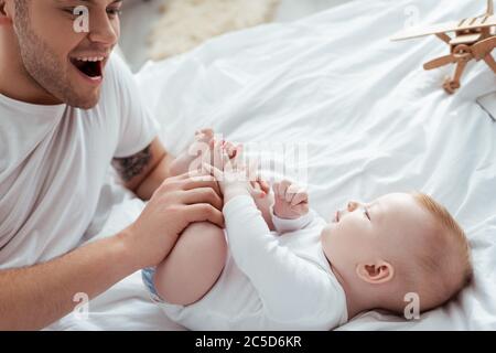 happy, excited father touching legs of adorable son lying in bed Stock Photo