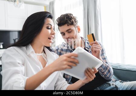 Selective focus of man holding credit card near excited girlfriend showing digital tablet at home Stock Photo