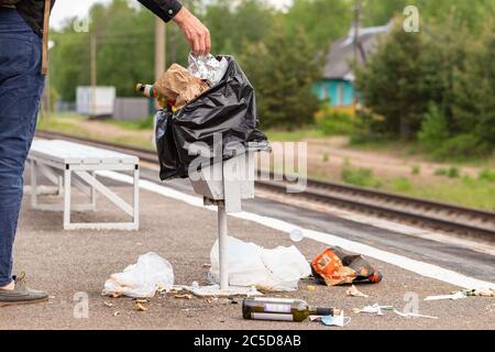 Hand of unknown man throws trash into overcrowded trash basket on a railway platform. Garbage bin. A pile of plastic waste trash on the floor. Stock Photo