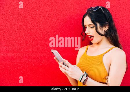 Woman looking at her phone with mouth wide open in red background Stock Photo