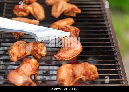 Grilled chicken wings on the grill with smoke, metal tongs to turning chicken wings. Close up, selective focus Stock Photo