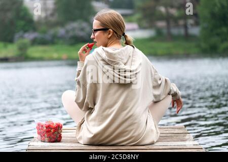 Young woman eating fresh berry fruit strawberries in plastic container, resting on a river wooden pier, back view. Summer time. The joy of country lif Stock Photo