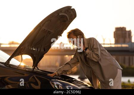 Helpless woman driver calling for help/assistance looking at broken down car, stopped at the roadside. Doesn't understand what happened. Sunset time. Stock Photo