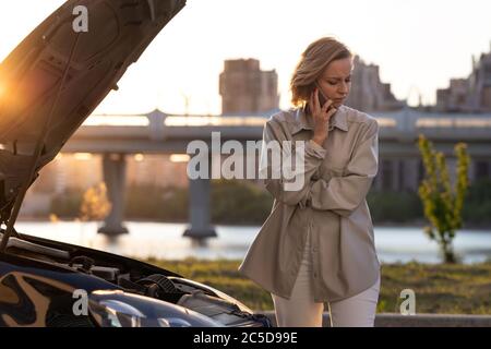 Helpless woman driver calling for help/assistance standing next to the broken down car, stopped at the roadside. Doesn't understand what happened. Stock Photo