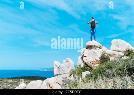 a young caucasian man, seen from behind, wearing jeans and a t-shirt and carrying a backpack, on the top of a rock formation, taking a photo of the se Stock Photo