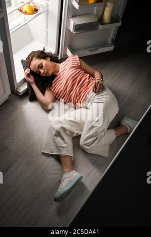 High angle view of young woman sleeping while lying near open refrigerator on floor at night Stock Photo