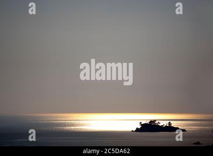 A lonely island in the light of the last rays of the sun that are reflected on the calm water of the sea at dusk. Stock Photo