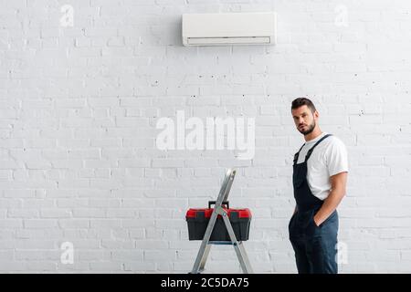 Handsome workman with hands in pockets of overalls looking at camera near toolbox on ladder and air conditioner Stock Photo