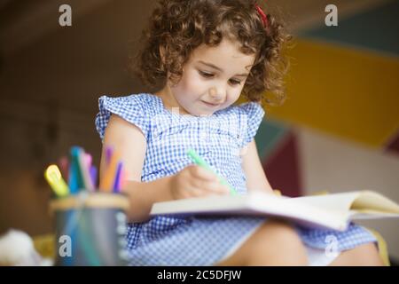 Beautiful little girl with dark curly hair in dress dreamily drawing in coloring book with felt-tip pens on sofa at home Stock Photo