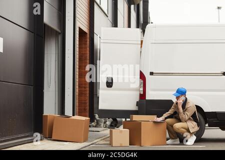 Young man talking on mobile phone and making delivery he sitting near van with boxes outdoors Stock Photo