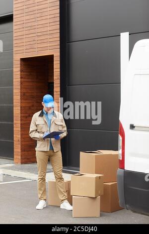 Young courier in uniform making notes in document he accepting parcels while standing outdoors near the warehouse Stock Photo