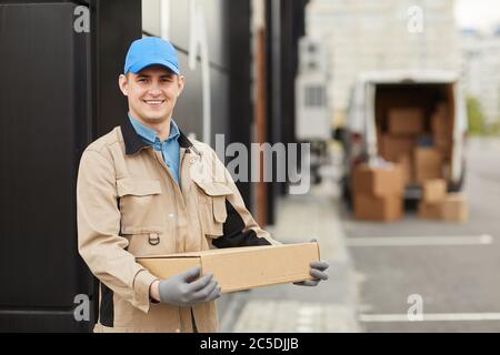 Portrait of young courier in uniform holding box and smiling at camera while standing outdoors Stock Photo