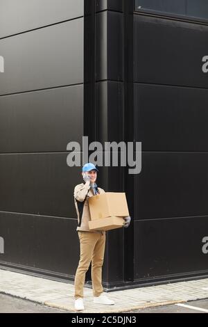 Young delivery person in uniform carrying boxes and talking on mobile phone while walking outdoors Stock Photo