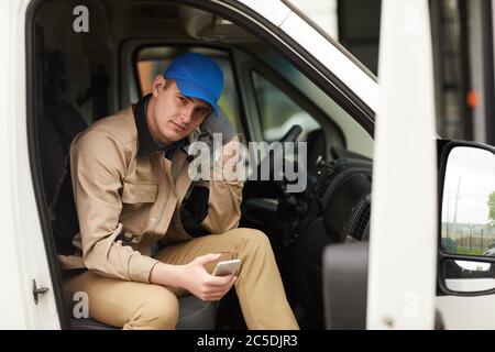 Portrait of delivery person in uniform holding mobile phone and looking at camera while sitting in the van Stock Photo