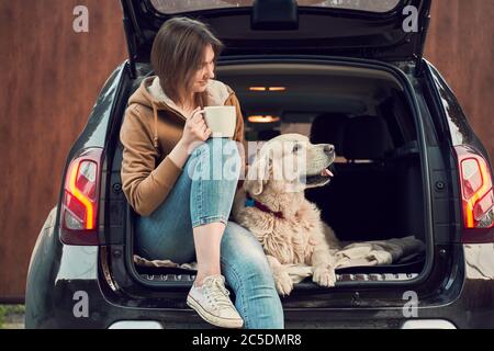 Young girl with mug in hand with dog sitting in open trunk of black car Stock Photo