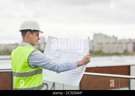 Serious engineer in work helmet examining the blueprint of new building while standing outdoors Stock Photo
