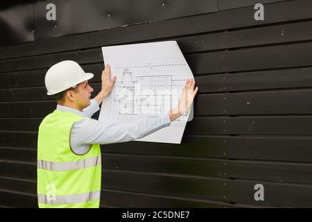 Young engineer in work helmet and reflective clothing examining the blueprint while standing outdoors Stock Photo