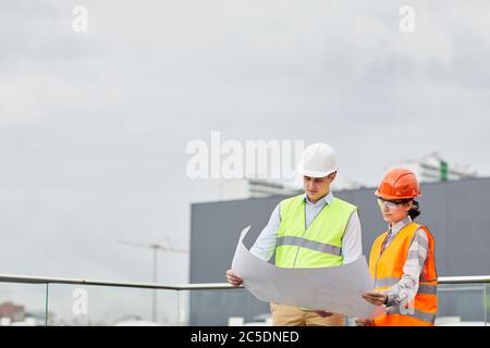 Two engineers in reflective clothing standing outdoors and examining the blueprint of new building together Stock Photo