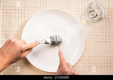 Minimal Easter composition. Hands cutting gray easter egg on white plate with fork and knife on linen tablecloth, glass of water Stock Photo