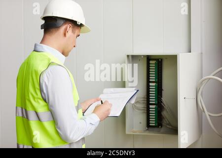 Rear view of engineer in reflective vest making notes in document he checking the data of electricity meter Stock Photo