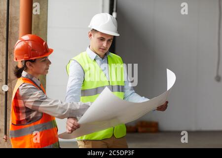 Two construction workers in work helmets examining the blueprint together while standing in empty building Stock Photo