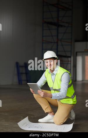 Portrait of young engineer in work helmet smiling at camera while working with blueprint and digital tablet in construction site Stock Photo