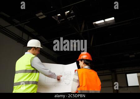 Rear view of two engineers in reflective vests looking at blueprint and discussing new construction project in team Stock Photo