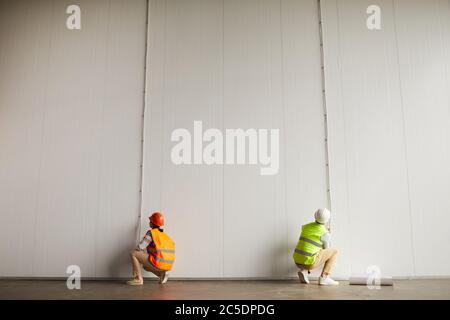 Rear view of two workers in reflective clothing working with wires on the white wall in the empty building on the construction site Stock Photo
