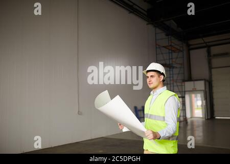 Mature engineer in reflective vest examining the plan of new building while standing in empty room Stock Photo