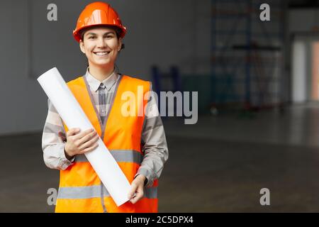 Portrait of young architect in work helmet and in reflective vest holding blueprint and smiling at camera while standing on construction site Stock Photo