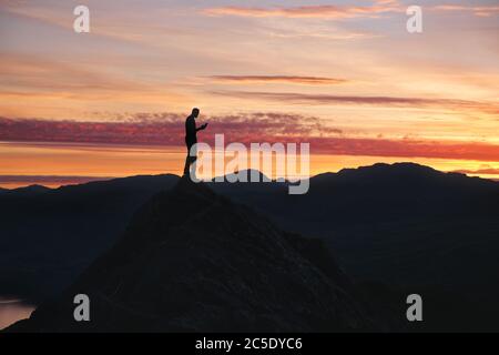 A silhouette of man using smart phone on the top of mountain against the backdrop of a stunning sunset sky. Scotland Stock Photo