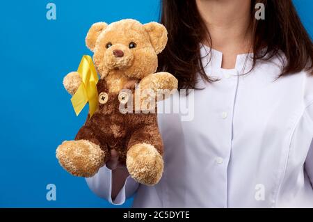 pediatrician in white coat holds a brown teddy bear with a yellow ribbon on a sweater, concept of the fight against childhood cancer, problem of suici Stock Photo