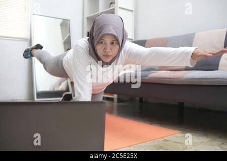 Asian muslim woman wearing hijab doing exercise at home while watching online video instruction on laptop, indoor home workout concept, keep healthy o Stock Photo
