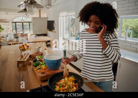 Happy african young woman talking on smartphone while preparing meal during covid-19 lockdown Stock Photo