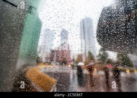 Rain droplets on a window with modern city architecture and people carrying umbrellas out-of-focus in the background. Stock Photo