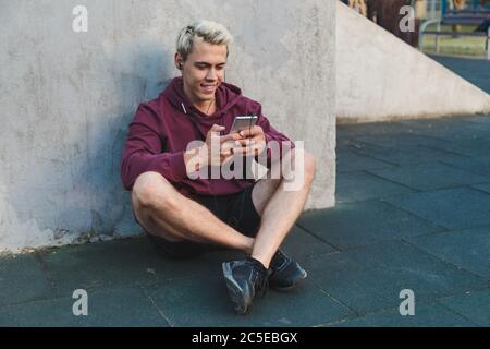Caucasian young man in sportswear sitting on the ground, resting after training and listening to music on headphones. Stock Photo