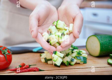 Hands of a young woman cut with a knife into cubes or slices of young zucchini cucumber on a wooden cutting board. Preparation of ingredients Stock Photo