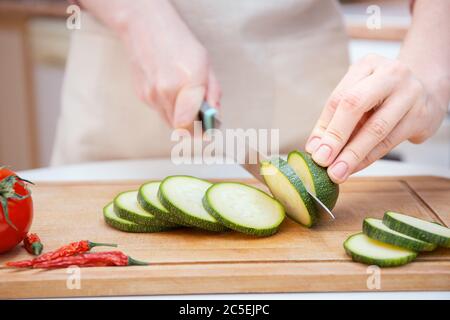 Close-up Hands of a young woman cut with a knife into slices or slices of young zucchini cucumber on a wooden cutting board. Preparation of ingredient Stock Photo