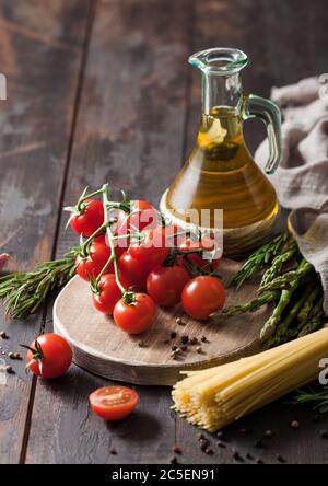 Olive oil with cherry tomatoes on round chopping board and with spaghetti pasta and rosemary and asparagus on wooden table background. Stock Photo