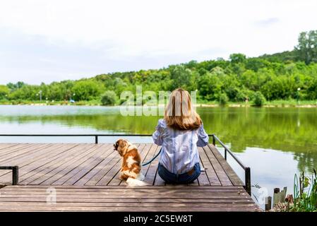 Rear view shot of woman wearing casual clothes while sitting on the pier next to her puppy. Copy space. Stock Photo
