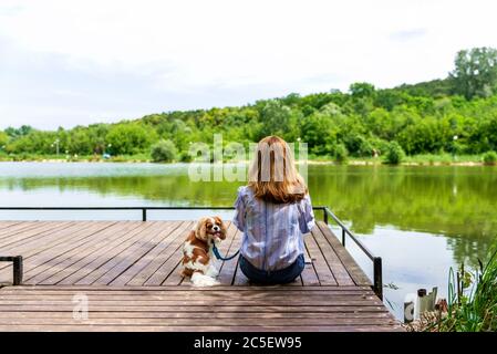 Rear view shot of woman wearing casual clothes while sitting on the pier next to her puppy. Copy space. Stock Photo