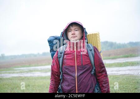 Guy tourist hiker  with backpack and travel mat, in a red jacket stands in the rain Stock Photo