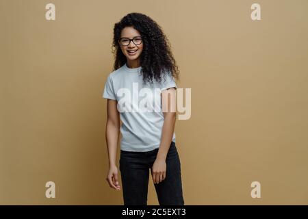 Smiling dark skinned young woman with glad face expression, wears optical glasses, white t shirt and jeans, poses carefree against beige background. P Stock Photo