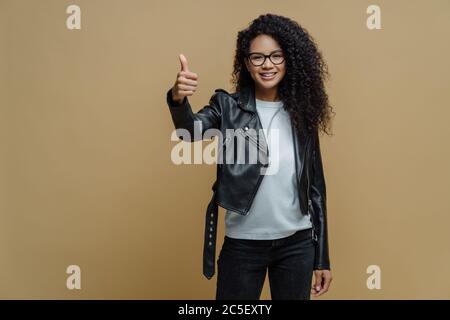 Confident cheerful African American woman shows thumb up in approval, gives recommendation or advice dressed in stylish leather jacket smiles positive