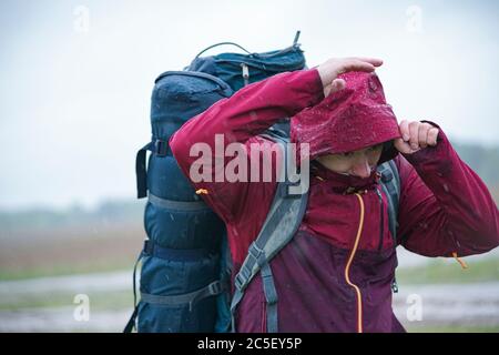 Guy tourist hiker  with backpack and travel mat, in a red jacket stands in the rain Stock Photo