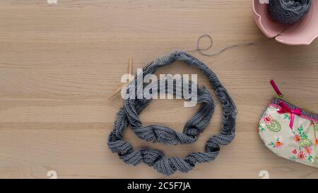 Flat lay of a gray blue colored knitwork on a wooden table, with a bowl holding the wool and a small purse with floral design. Stock Photo