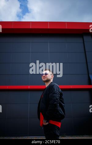 A young man with a beard in glasses in a red fashionable t-shirt with a jacket poses near a gray building. Attractive guy resting in the city on a sum Stock Photo