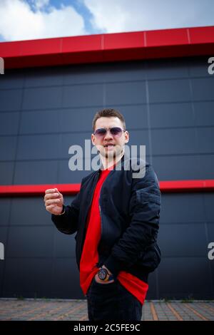 A young man with a beard in glasses in a red fashionable t-shirt with a jacket poses near a gray building. Attractive guy resting in the city on a sum Stock Photo