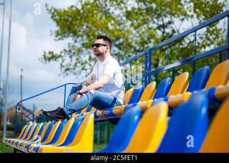 A guy in a white T-shirt sits in a stadium on the chairs for the fans alone. Stock Photo